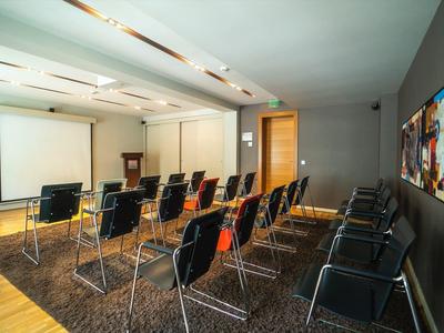 Nowoczesna sala konferencyjna z rzędami krzeseł skierowanymi na ekran projekcyjny, neutralne kolory i duży obraz.