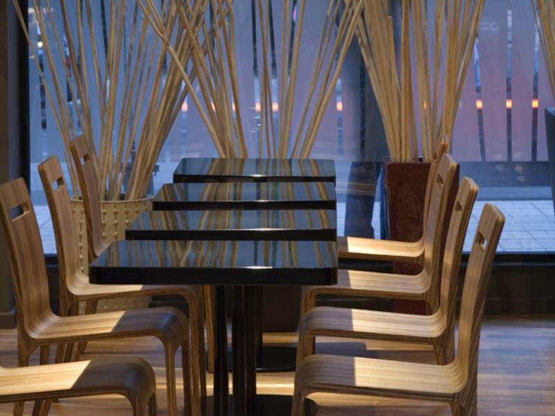 Nowoczesna jadalnia z wieloma stołami, krzesłami i dekoracyjnymi drewnianymi patyczkami przed dużymi oknami.