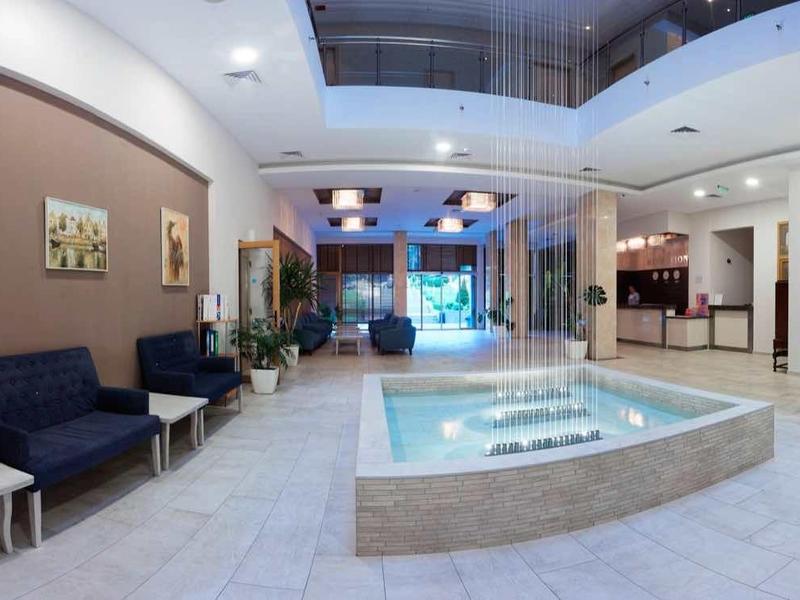 Spacious hotel lobby with light walls, armchairs, atrium with glass ceiling, and central fountain.