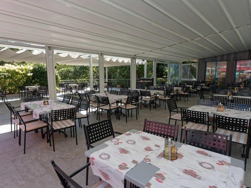 Large terrace with numerous tables and chairs under a roof, surrounded by green vegetation.
