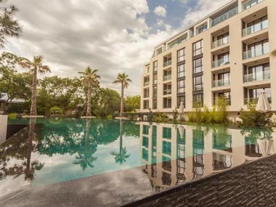 Moderner Pool vor mehrstöckigem Gebäude, Palmen und bewölktem Himmel, spiegelnde Wasseroberfläche.