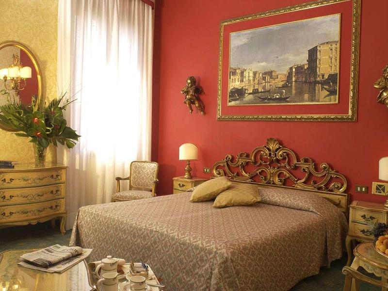 Elegant hotel room with red wall, double bed, and gold-accented furniture.