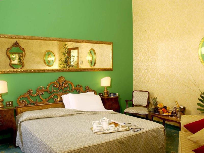 Elegant hotel room with green and yellow decor, large bed, and classic furniture.