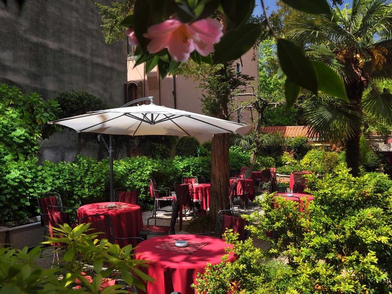 Sunny outdoor area with tables, red tablecloths, and a white umbrella among green plants.