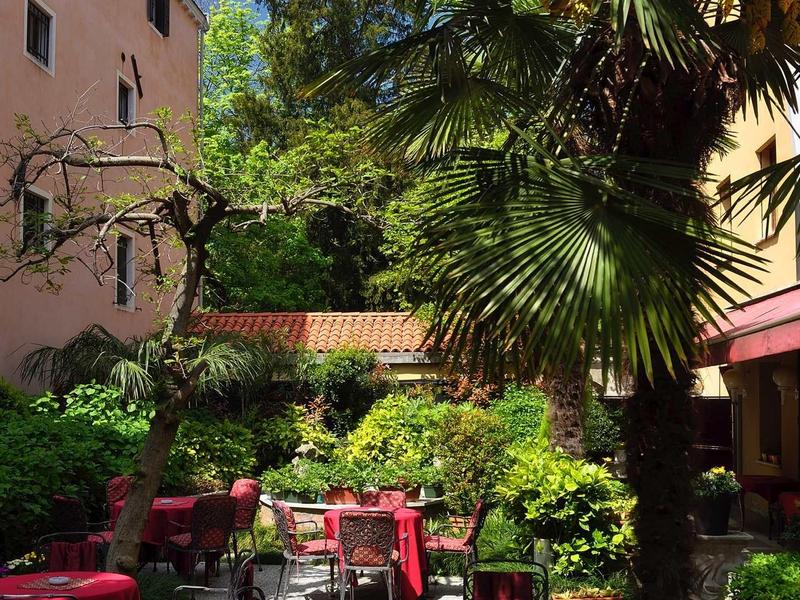 Outdoor area with tables and chairs surrounded by lush plants and palm trees beside buildings.