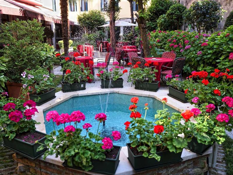 Lively garden with small octagonal water feature surrounded by flowers and red chairs.