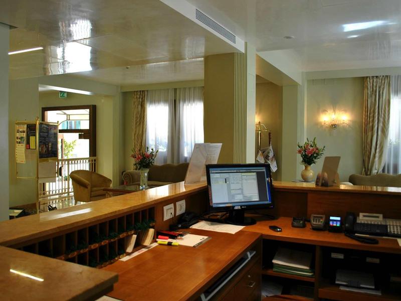 Modern hotel lobby with reception desk, computer setup, and chairs in warm lighting.