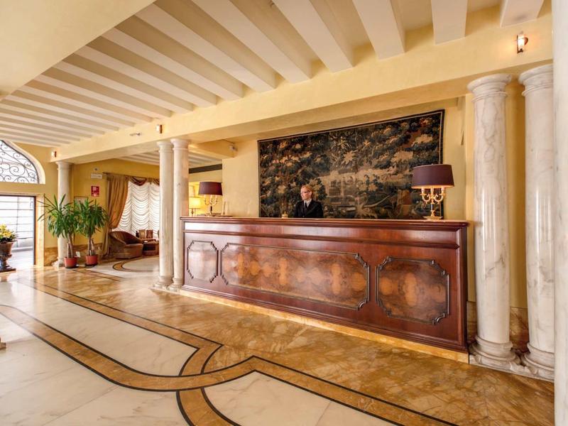 Elegant hotel lobby with wooden reception desk, marble floor, and columns under bright ceiling beams.