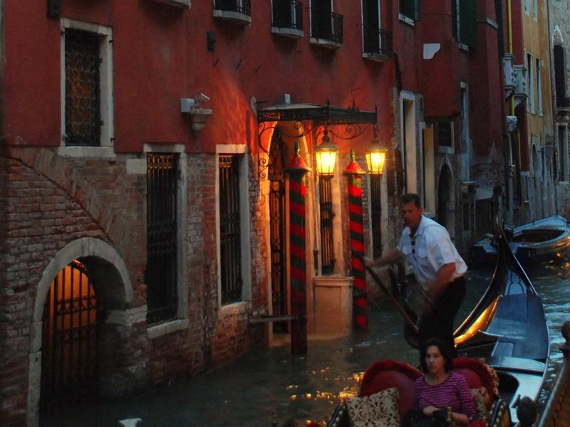 Wieczorna scena z gondolą na weneckim kanale przed czerwonym budynkiem.