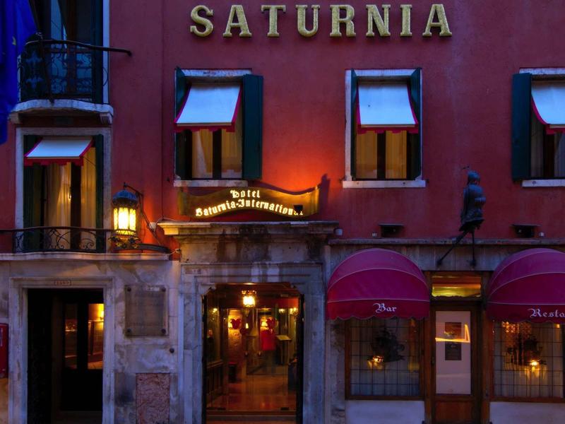 Wieczorny widok na oświetlone wejście do hotelu Saturnia z czerwoną elewacją i oknami.