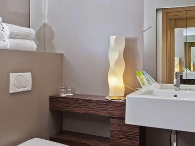 Modernes Badezimmer mit Wand-WC, weißem Waschbecken, Holzoptikablage und leuchtender Dekorlampe.
