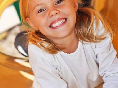 Lächendes blondes Mädchen mit Zöpfen sitzt in gelbem Spieltunnel, trägt weißen Pullover.