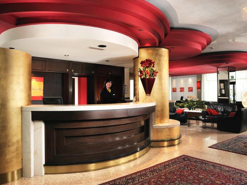 Hotelrezeption mit rundem Tresen, roten Deckenakzenten und goldenen Säulen in elegantem Stil.