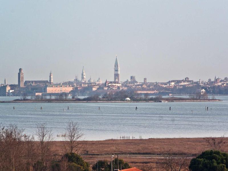 Stadtpanorama mit historischen Gebäuden und Turm am Ufer eines ruhigen Gewässers bei klarem Himmel.