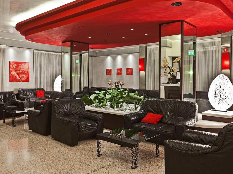 Moderne Lobby mit schwarzen Ledersesseln, roten Akzenten und großen Pflanzen auf mittigem Tisch.