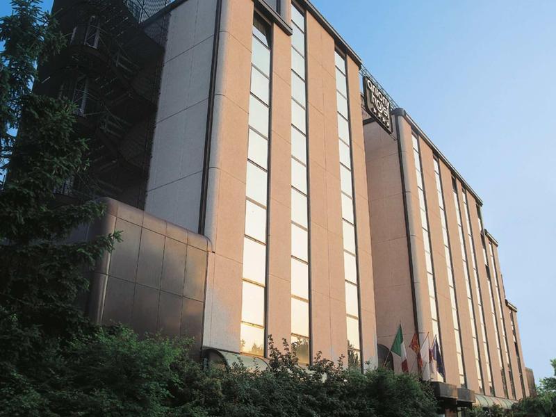 Ein modernes, mehrstöckiges Gebäude mit großen Fenstern, umgeben von Grün und blauem Himmel.