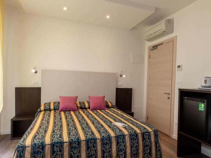 Hell eingerichtetes Schlafzimmer mit Doppelbett, bunten Kissen und Seitentischen an weißen Wänden.