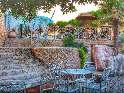 Terrasse mit Metalltischen und -stühlen, Treppe aus Naturstein, umgeben von Bäumen und Pflanzen.