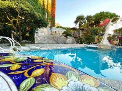 Bunt verzierter Tisch neben einem klaren blauen Pool mit Garten und Statue im Hintergrund.