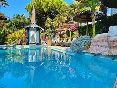 Klares blaues Poolwasser vor einem tropischen Garten mit Sonnenschirmen und einer kleinen Säule.