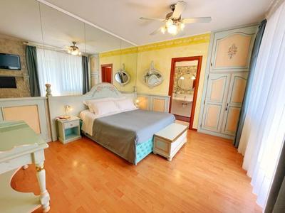 Helles Schlafzimmer mit Doppelbett, Holzfußboden, Deckenventilator und großen Fenstern mit weißen Vorhängen.