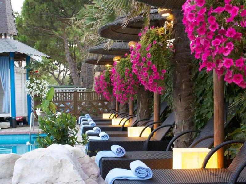 Reihe von Liegestühlen mit blauen Handtüchern, umgeben von pinken Blumen und grünen Pflanzen.