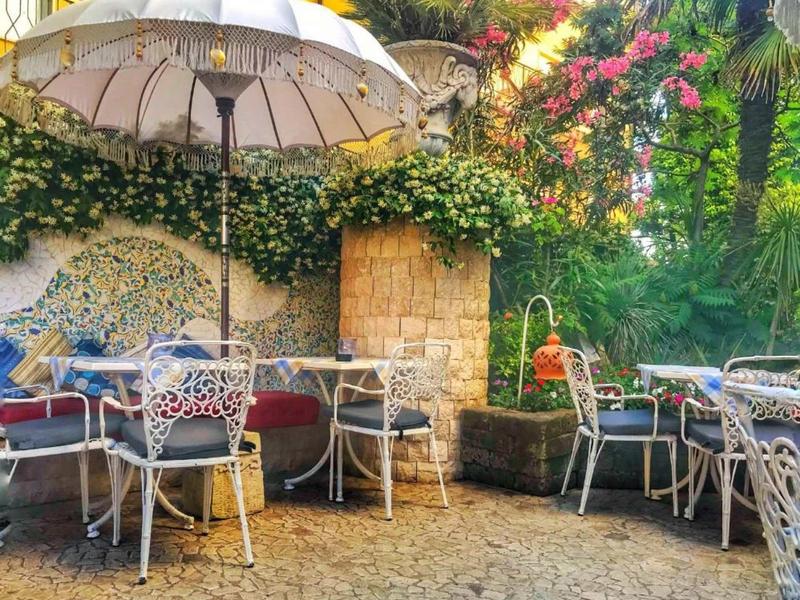Gemütlicher Gartensitzplatz mit Metallstühlen, Tischen, Sonnenschirm und üppigem Blumenbewuchs.
