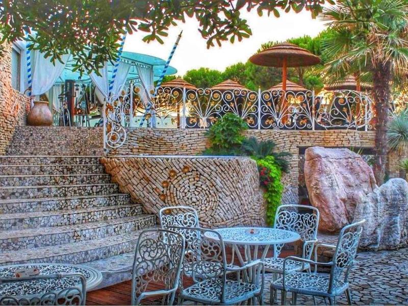 Terrasse mit Metalltischen und -stühlen, Treppe aus Naturstein, umgeben von Bäumen und Pflanzen.