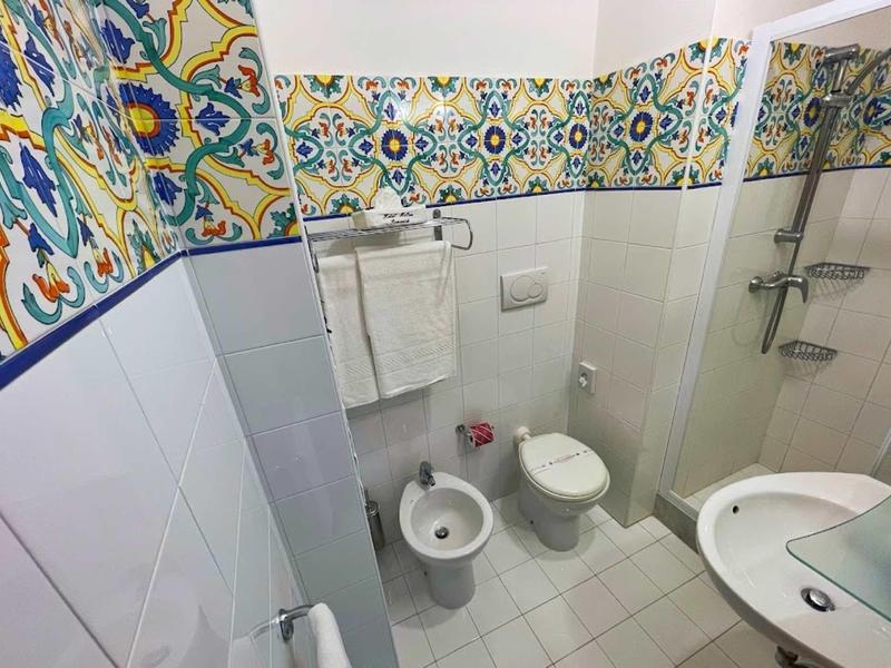 Kleines Bad mit weißer Kloschüssel, Bidet, Duschkabine, Waschbecken und farbiger Fliesenbordüre.