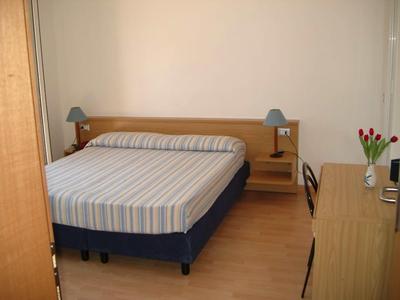 Hotelový pokoj s manželskou postelí, dvěma nočními stolky s lampami, psacím stolem a váza s květinami.