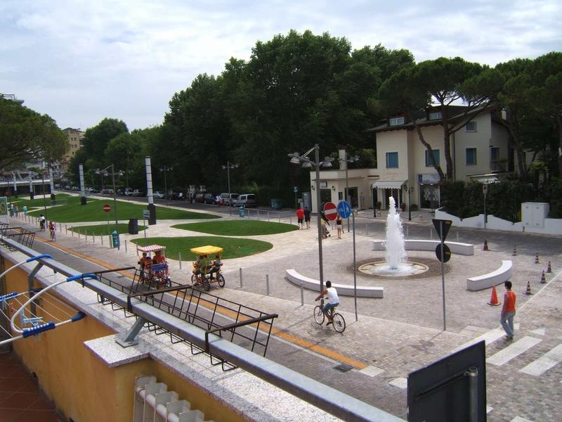 Veřejné náměstí s fontánou a lidmi v městském prostředí obklopeném stromy.