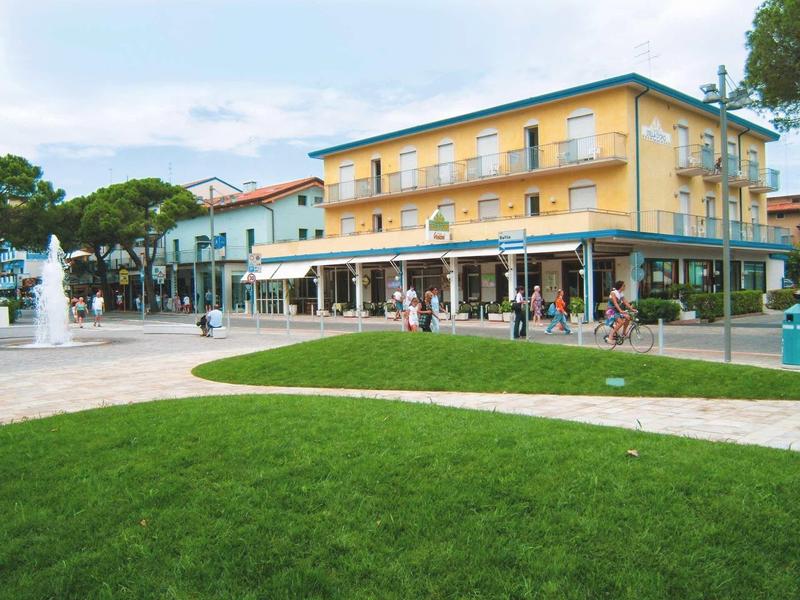 Světle žlutá hotelová budova s verandou na rušném náměstí a zeleným trávníkem v popředí.