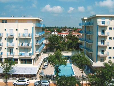Two modern hotels with a swimming pool between them, surrounded by trees and cars.