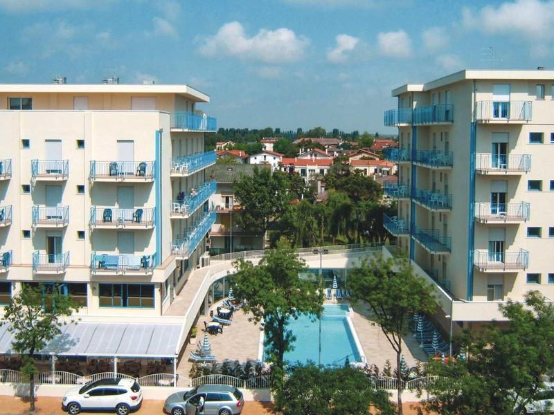 Two modern hotels with a swimming pool between them, surrounded by trees and cars.