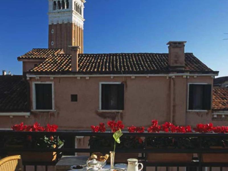 Stůl prostřený na balkoně s červenými květinami a věží v pozadí.