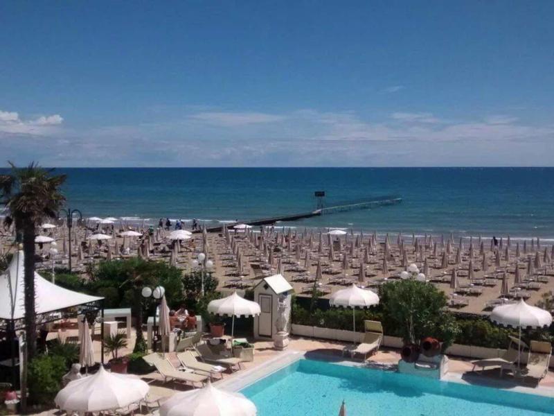 View of a busy beach with umbrellas, pool, and sea in the background.