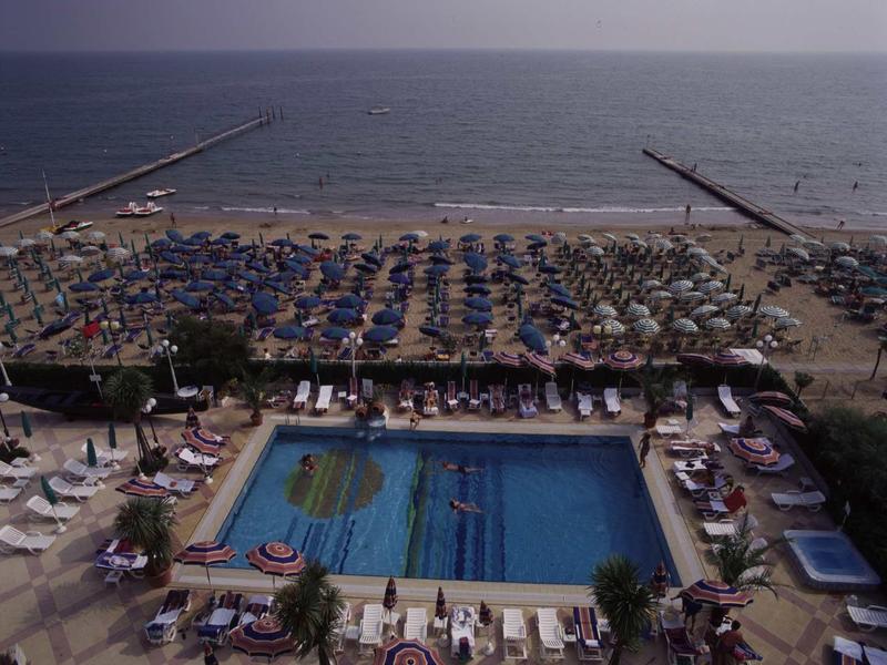 Overview of a busy beach with blue umbrellas and a swimming pool in the foreground.