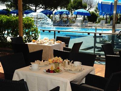 Restaurantterrasse mit gedeckten Tischen, schwarzen Stühlen, blauem Pool und Sonnenschirmen im Hintergrund.