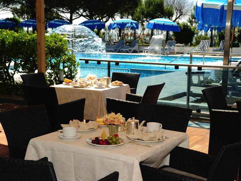 Tavolo con tazze di caffè e pasticcini davanti a una piscina con ombrelloni blu.