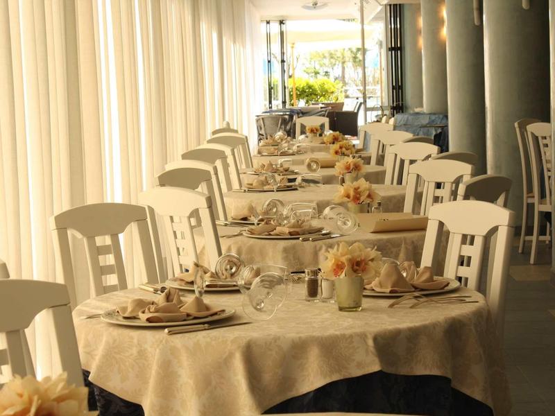 Ristorante elegante con una lunga tavola apparecchiata e sedie bianche accanto a grandi finestre.
