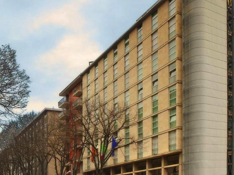 Moderní hotelová budova se šesti patry oken na ulici lemované stromy.