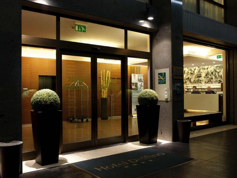 Vstup do hotelu v noci se skleněnými dveřmi a dekorativními rostlinami v černých květináčích.