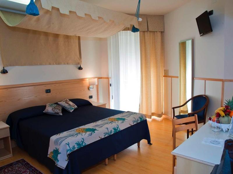 Helles Schlafzimmer mit Doppelbett, Holzboden, Sessel, Schreibtisch und Balkon mit Gardinen.