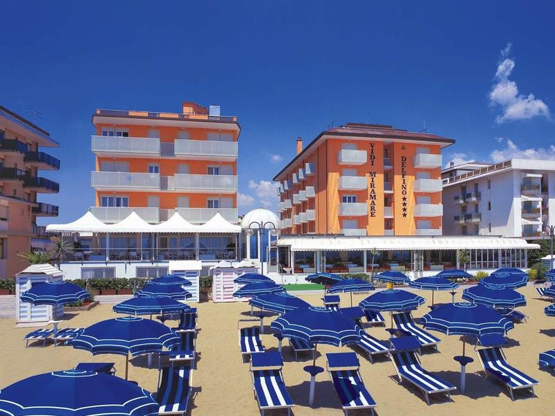 Strand mit blauen Sonnenliegen und Schirmen vor orange-weißen Mehrfamilienhäusern unter blauem Himmel.