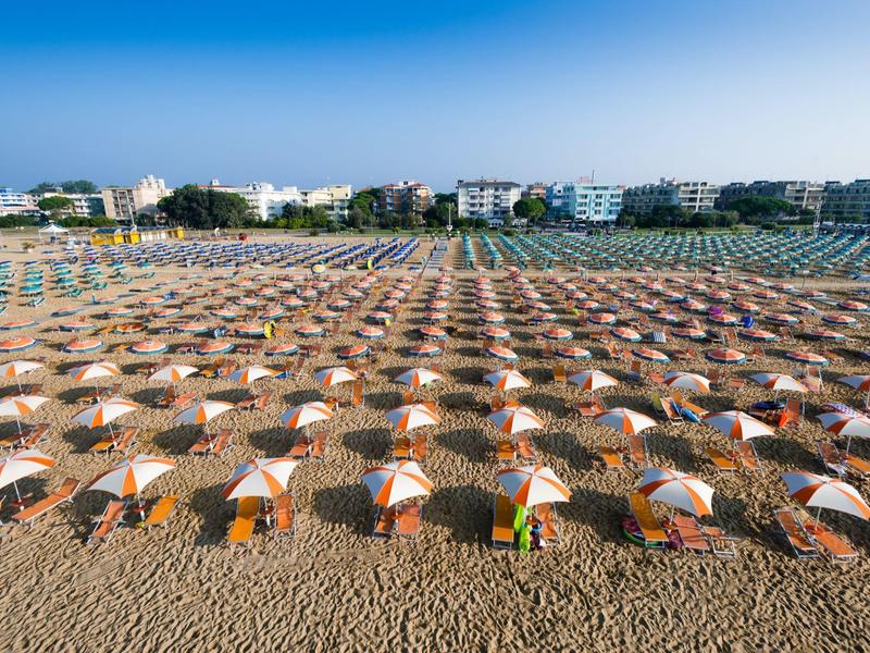 Rows of beach umbrellas on sand with buildings in the background under clear sky.