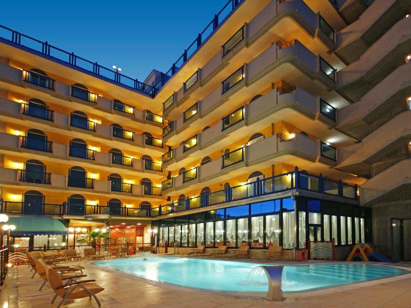 Außenansicht eines mehrstöckigen Hotels mit beleuchtetem Pool und Sitzbereich am Abend.