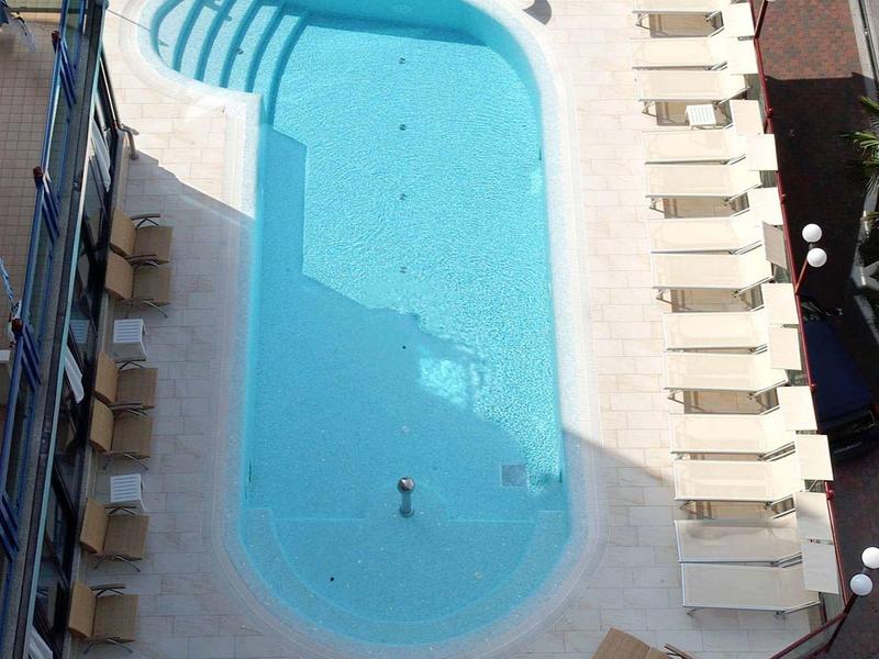 Vogelperspektive eines rechteckigen Pools mit Liegestühlen und Tischen drumherum.