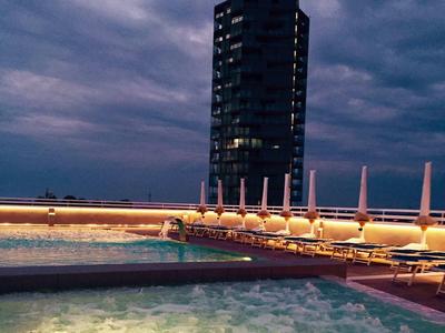 Beleuchteter Außenpool mit Whirlpool bei Dämmerung und modernem Hochhaus im Hintergrund.