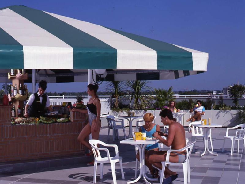 Frau in Badeanzug am Tresen eines Außen-Cafés, weitere Gäste sitzen an weißen Tischen unter grün-weißem Sonnendach.