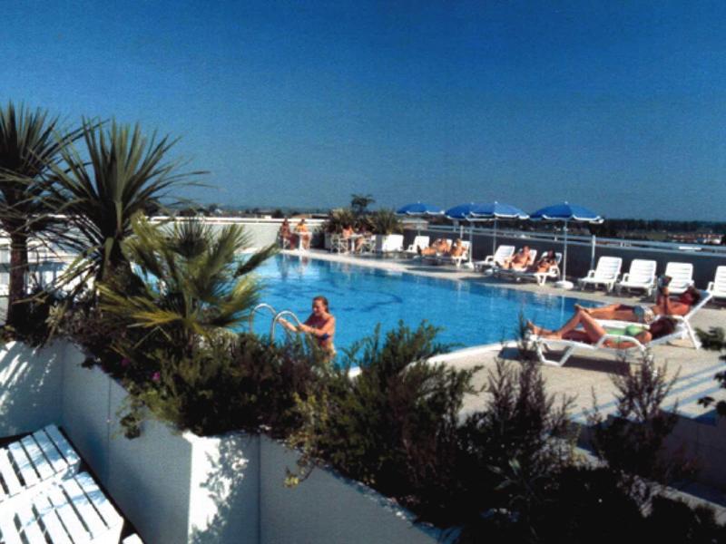 Schwimmbad mit Liegestühlen, Pflanzen und Meer im Hintergrund bei klarem Himmel.